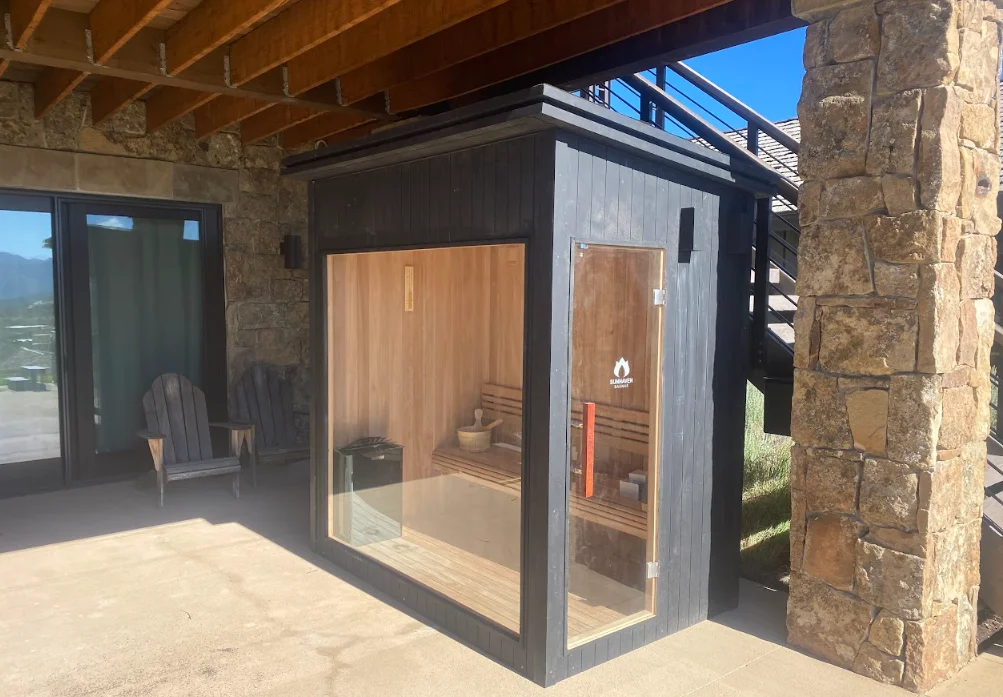 Cedar sauna with glass door and window, black frame, installed on patio next to stone chimney