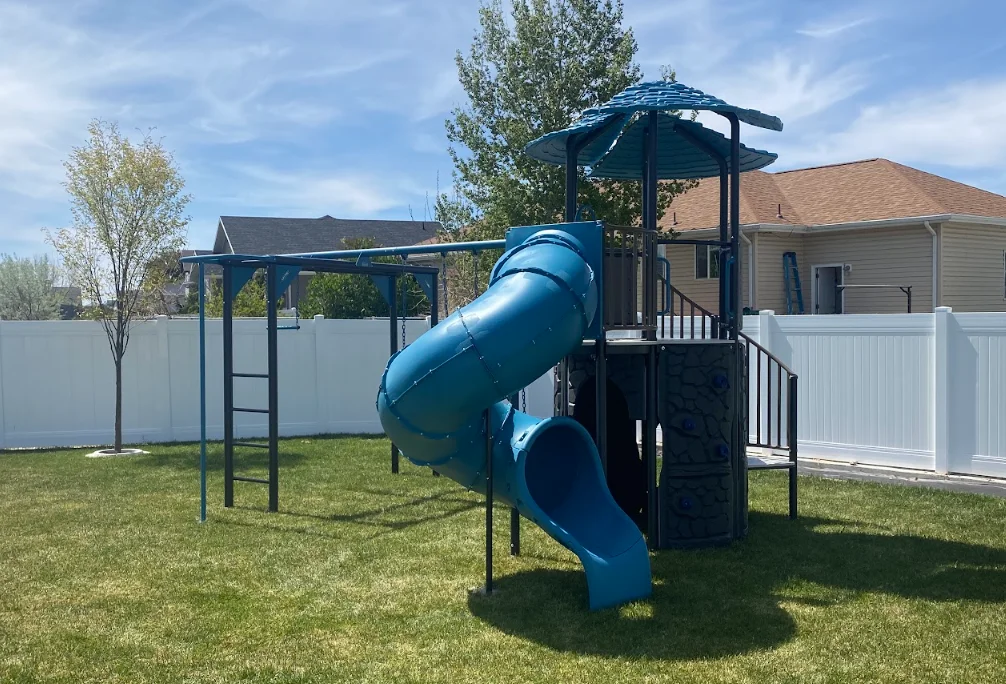 Blue playset with spiral slide, climbing ladder, and pergola frame installed in fenced backyard