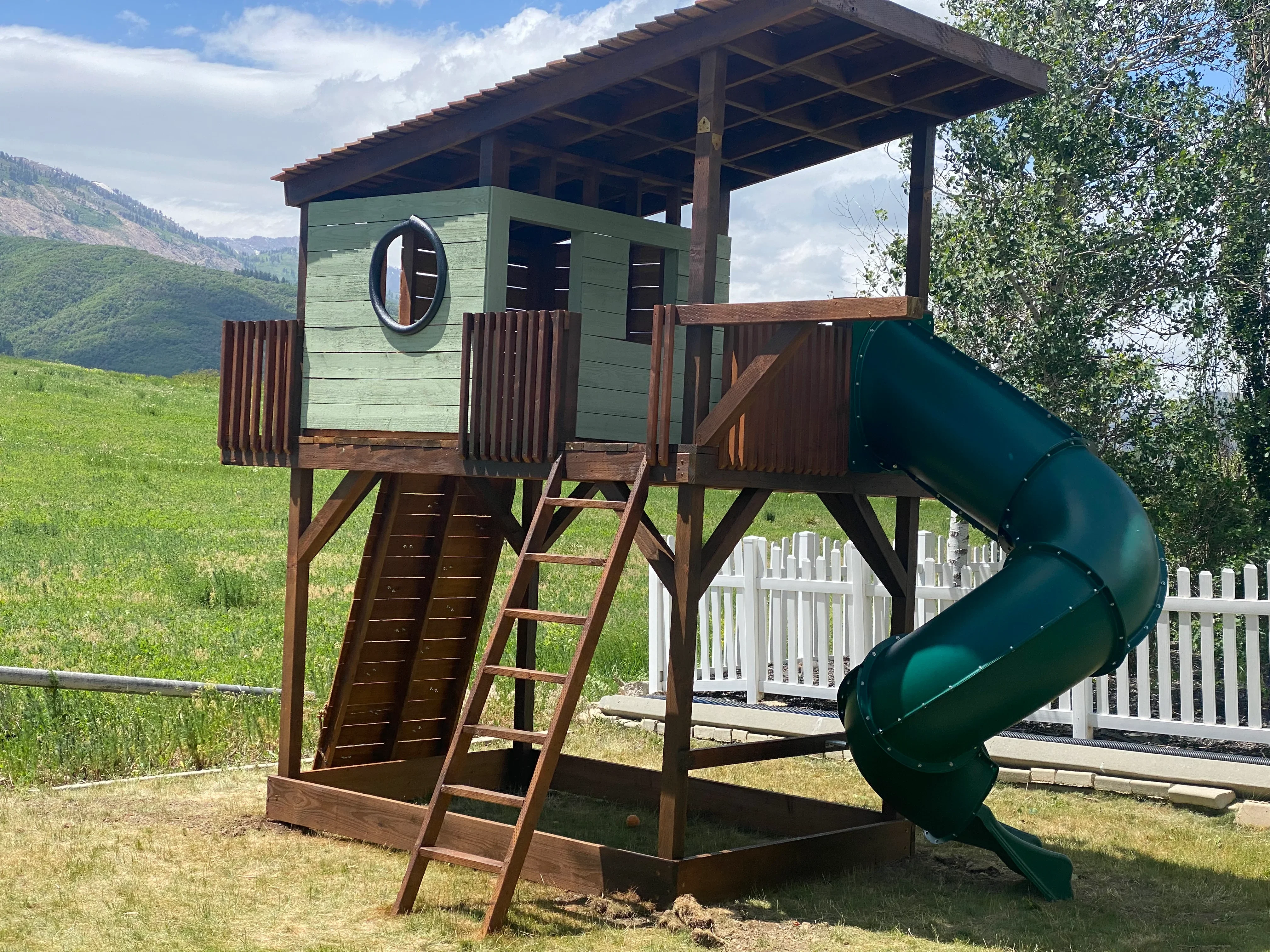 Elevated wooden playhouse with green cabin walls, spiral slide, and timber frame construction