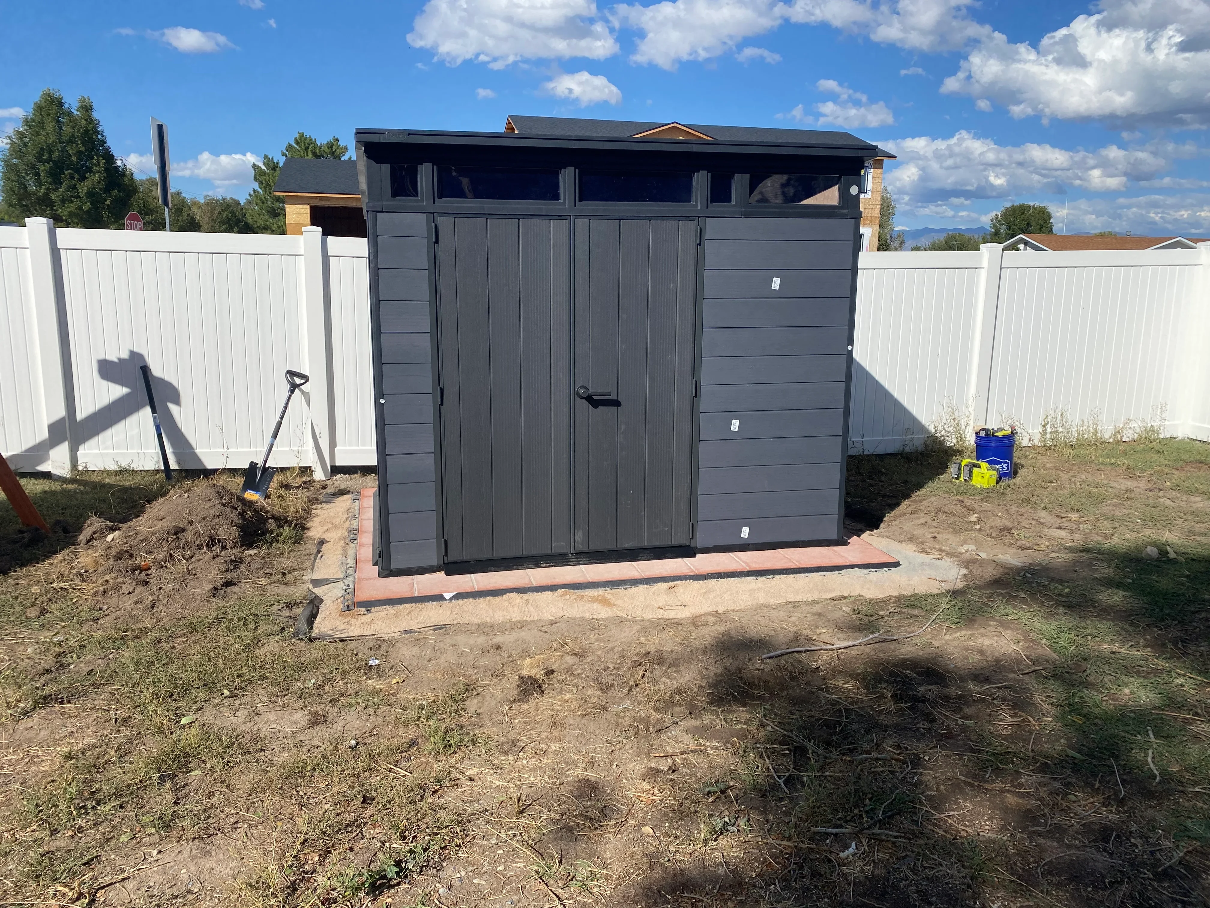 Dark gray metal storage shed with double doors on brick foundation, white vinyl fencing on sides