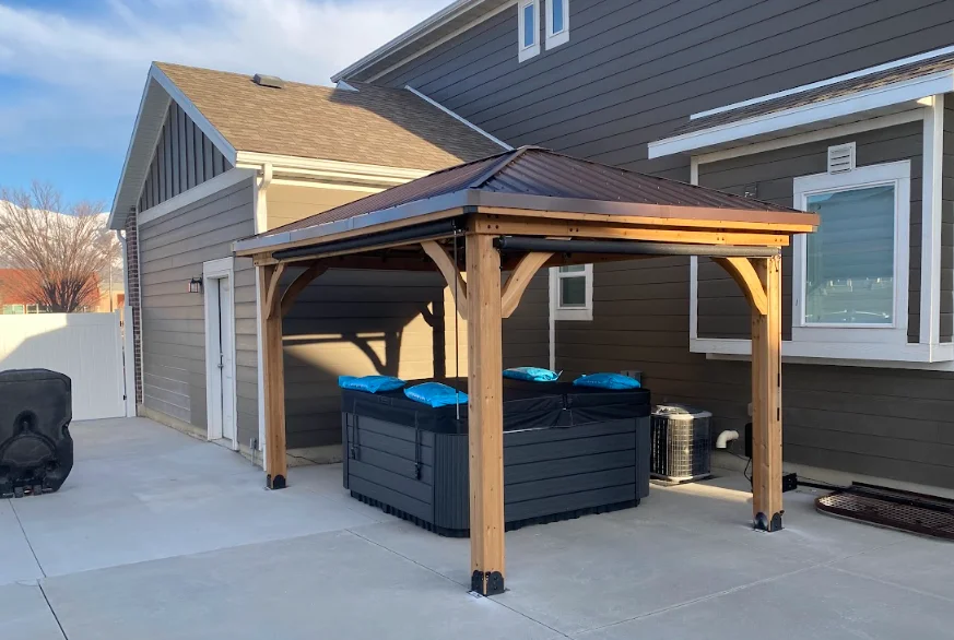 Wooden gazebo with metal roof covering two hot tubs on concrete patio
