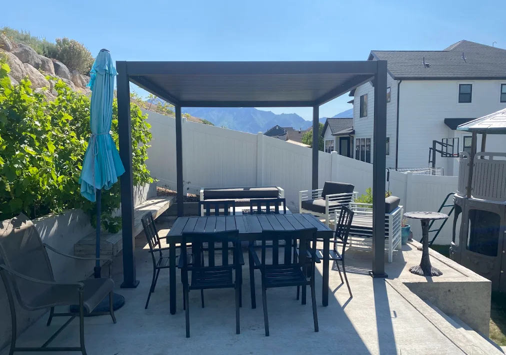 Black aluminum pergola with slatted roof over concrete patio and dining furniture