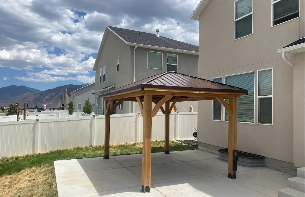 Wooden gazebo with metal roof on concrete patio beside residential home