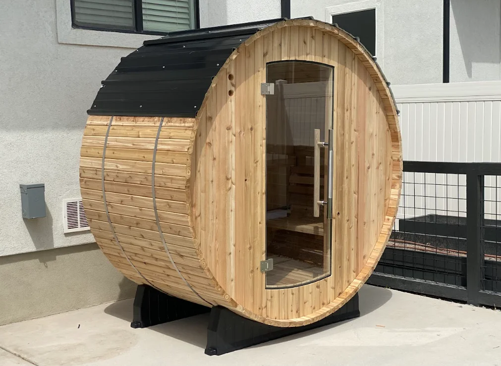 Cedar barrel sauna with black metal roof and glass door installed on concrete patio