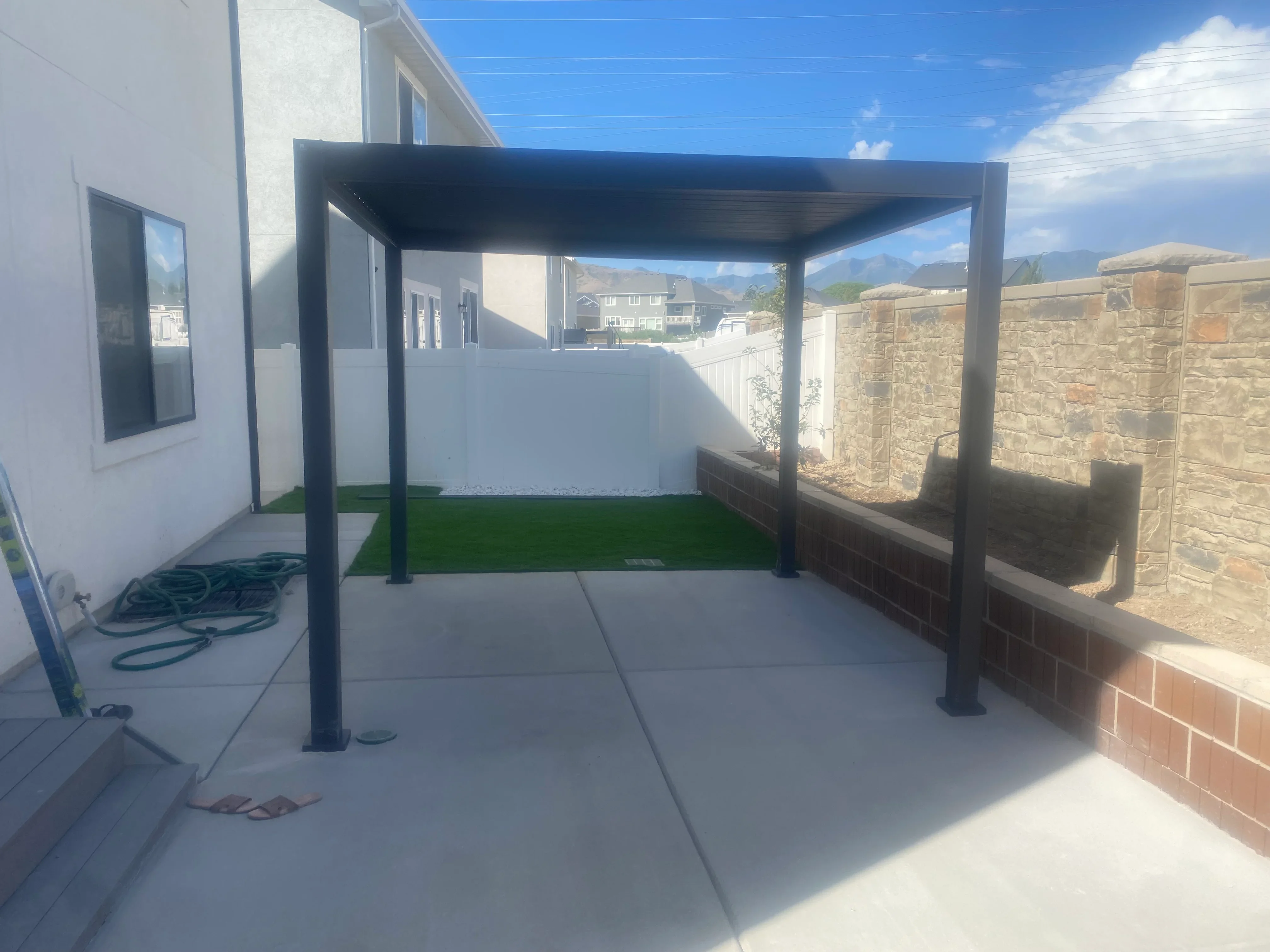 Black metal pergola over concrete patio with artificial turf and stone retaining wall