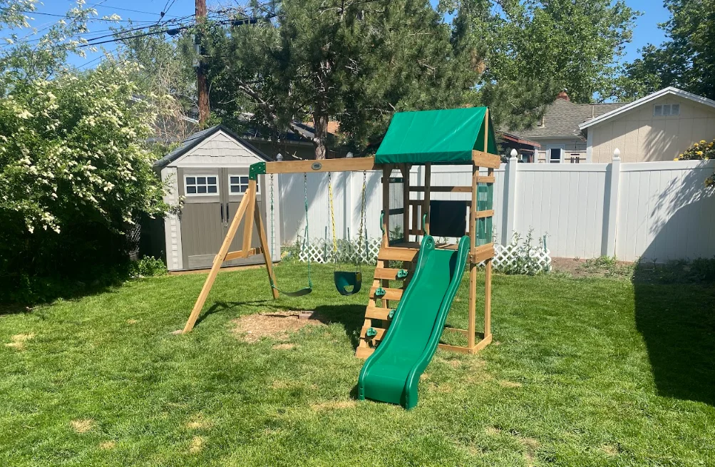 Wooden playset with green slide, swing, and canopy installed in residential backyard