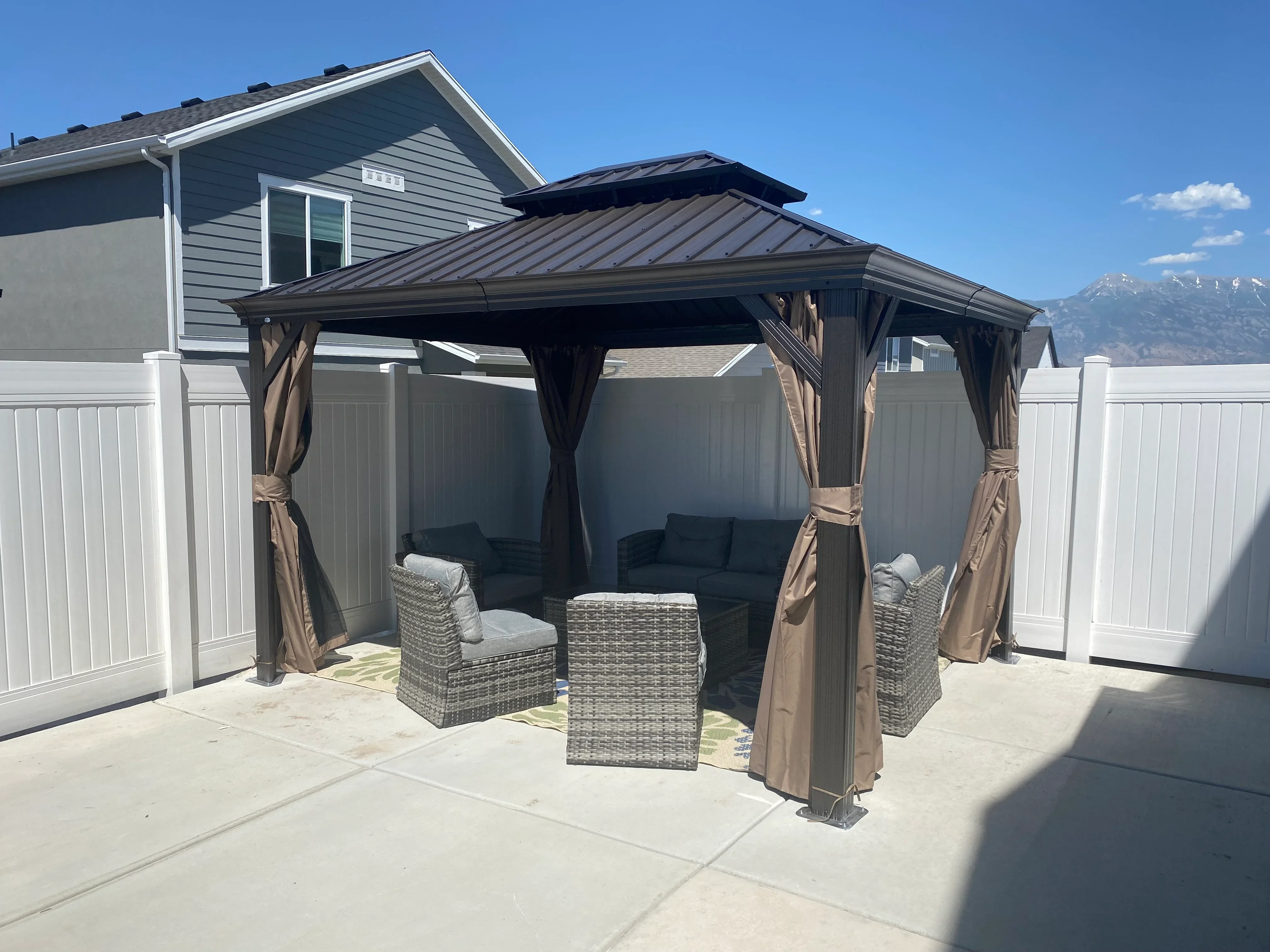 Black hardtop gazebo with metal roof, tan curtains, and wicker furniture on concrete patio