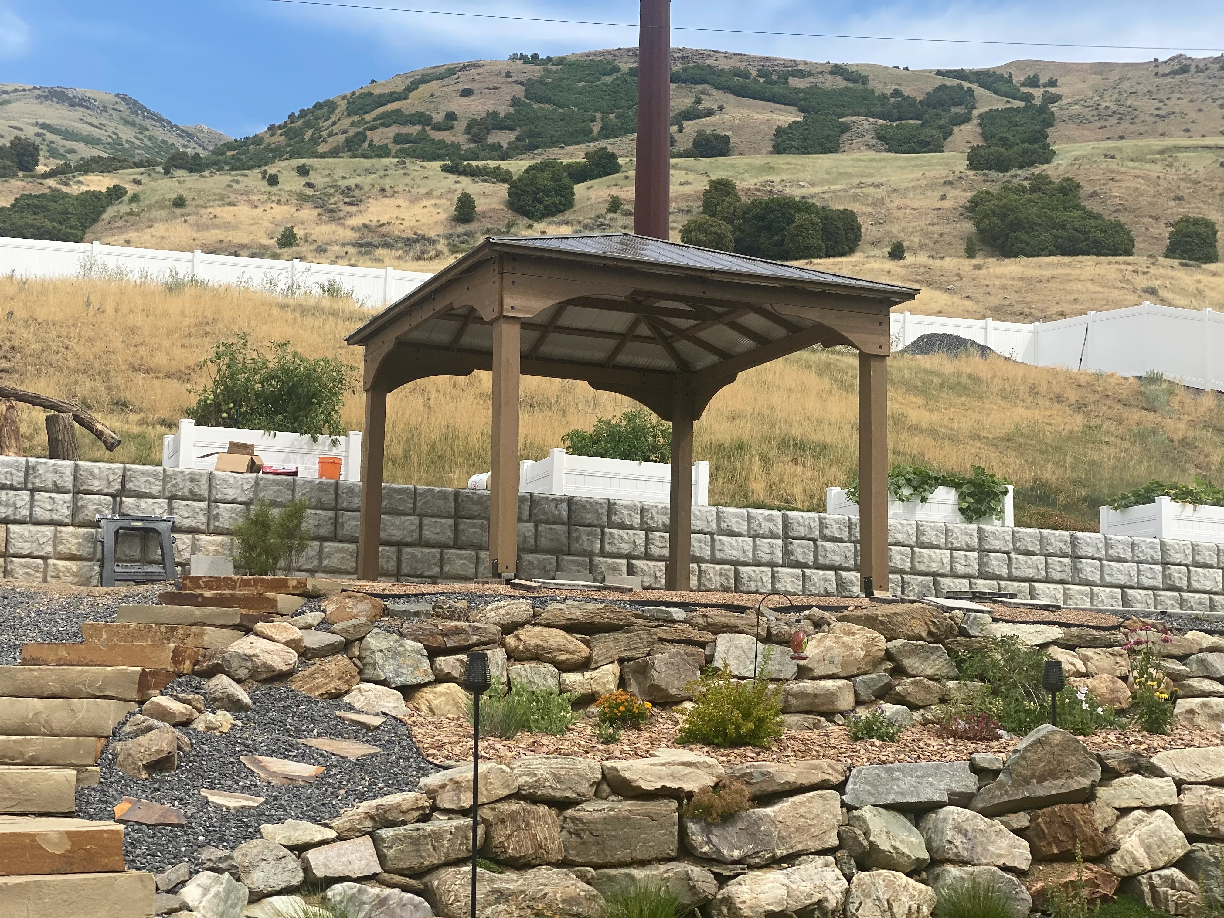 Stacked stone retaining wall with timber pergola and metal roof, gravel pathways, and landscape plantings