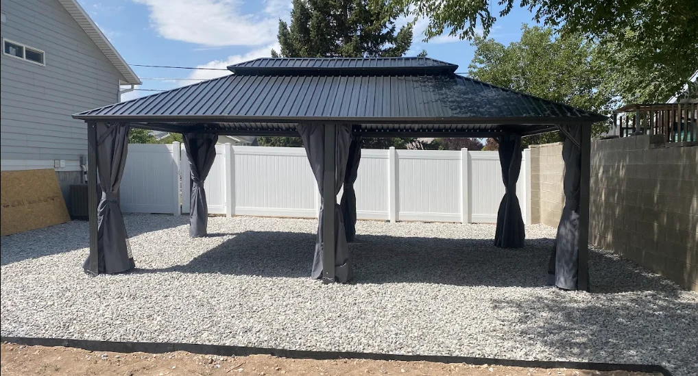 Black metal gazebo with peaked roof and gray curtains on gravel base in backyard