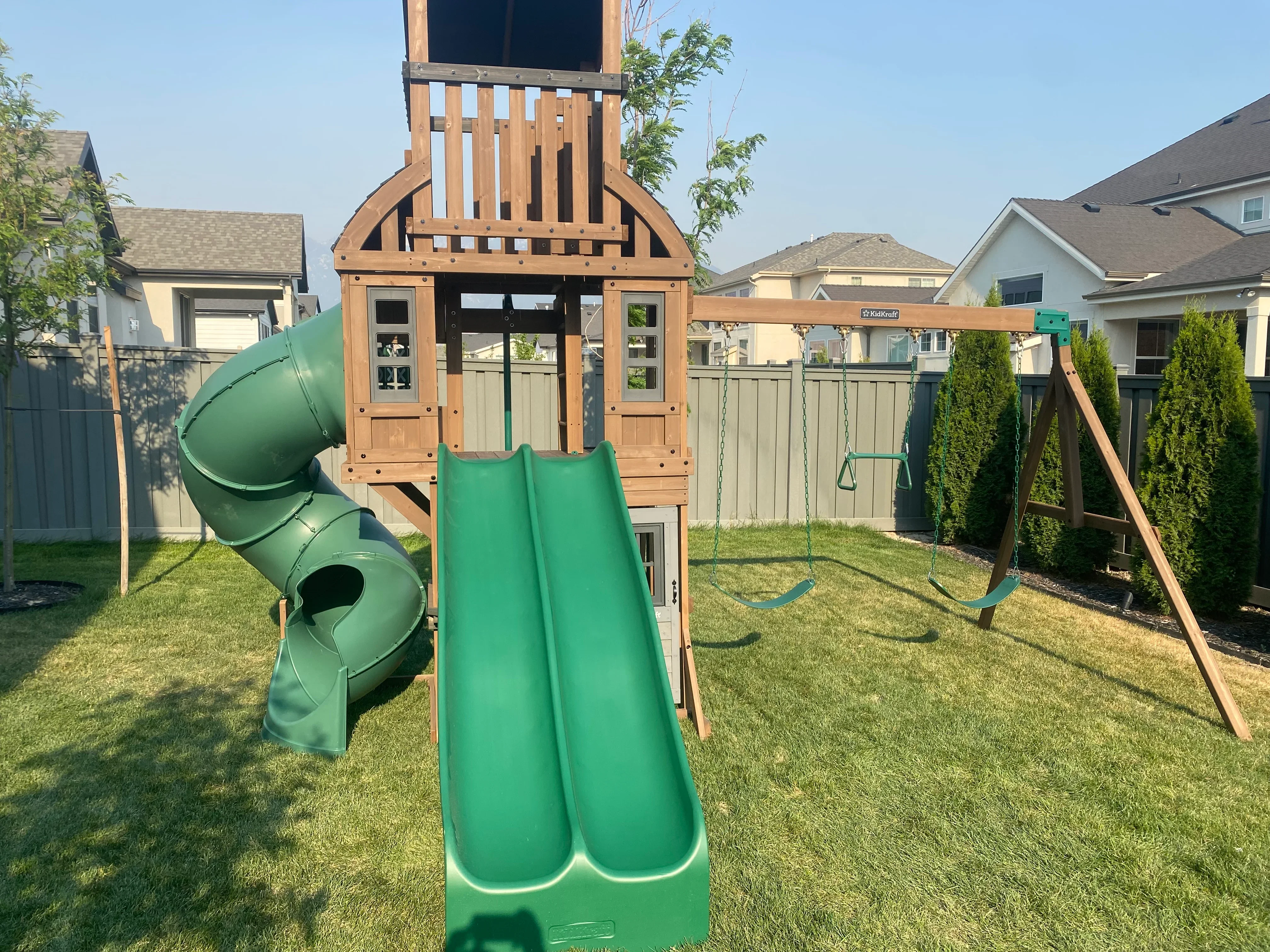 Cedar wood playground tower with green slides, tube slide, and swing set installed in residential backyard