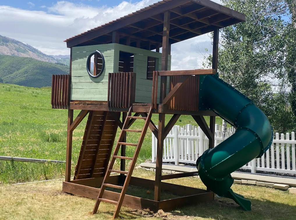 Elevated wooden playset with green cabin tower, spiral slide, and timber frame roof
