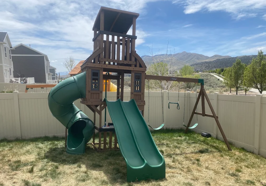 Wooden playset with green slides, swings, and monkey bars in fenced backyard