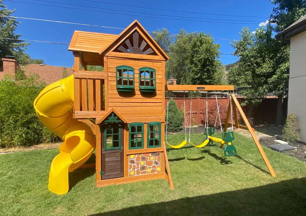 Two-story wooden playset with yellow spiral slide, swings, and sandbox on grass lawn