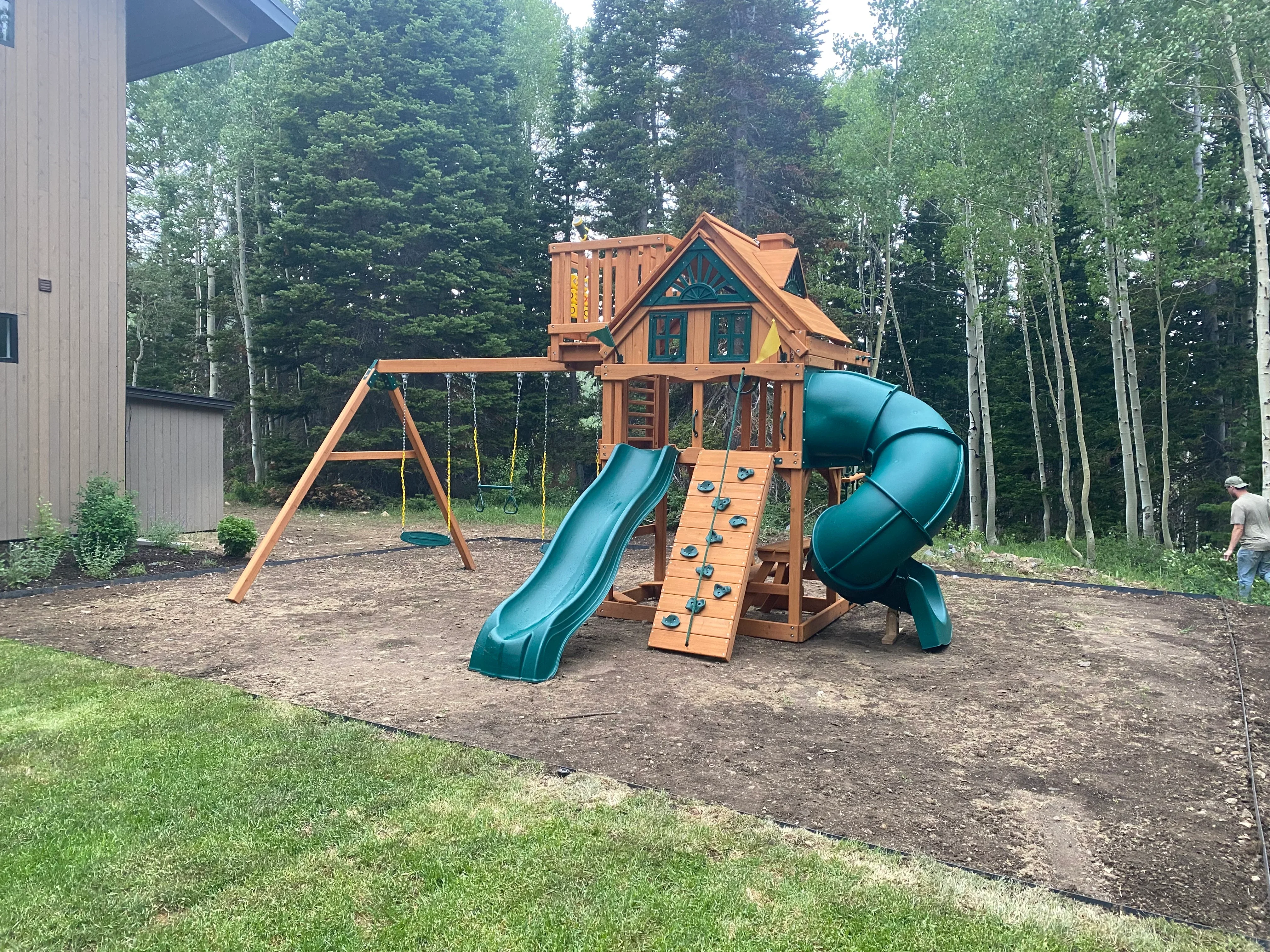 Wooden playground structure with turret, teal slides, climbing wall, and swings in backyard setting