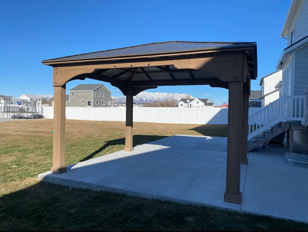 Wood and metal pergola with black roof panels over concrete patio in backyard