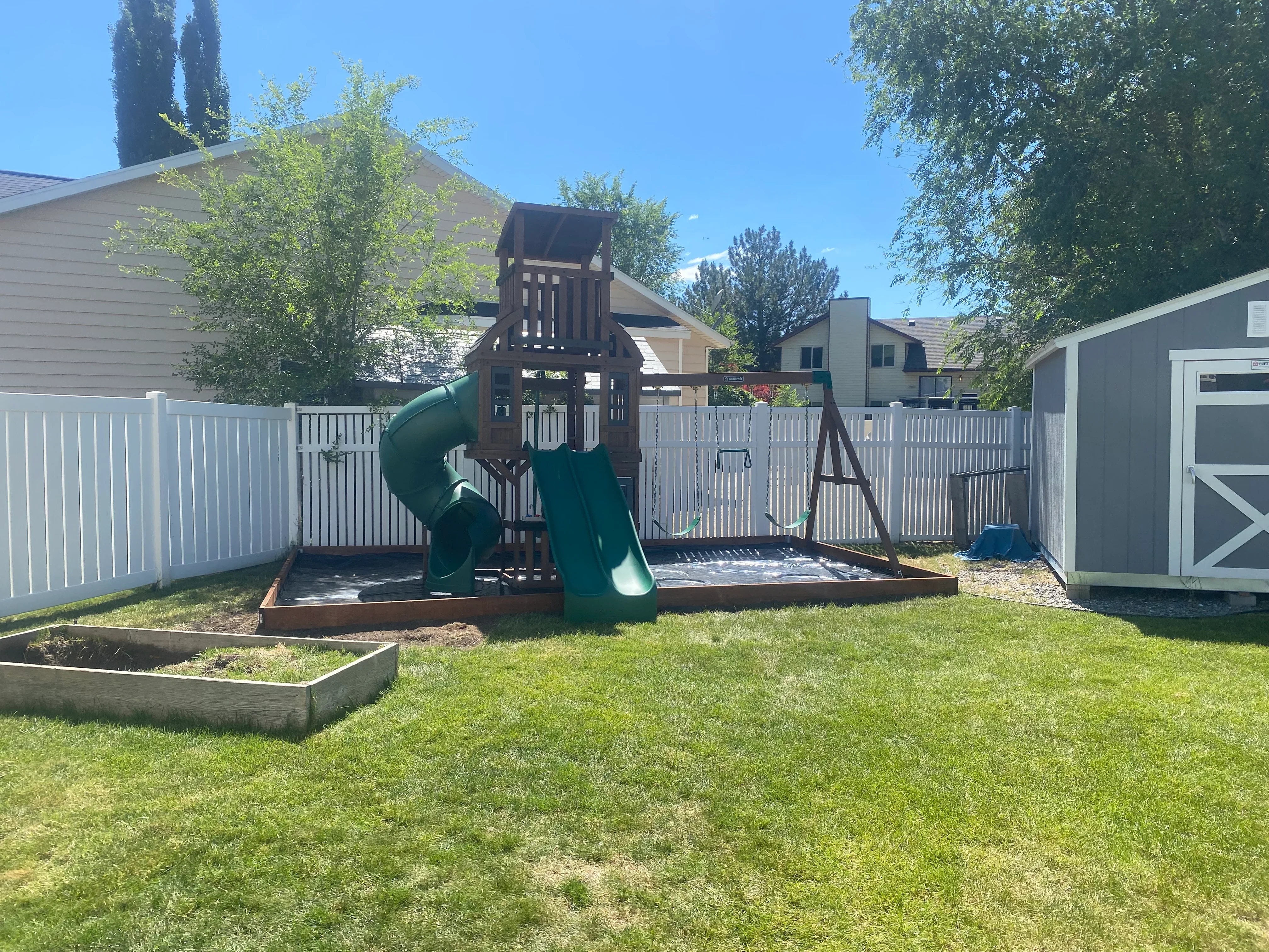 Wooden play tower with green spiral slide, sandbox, and white vinyl fence in residential backyard