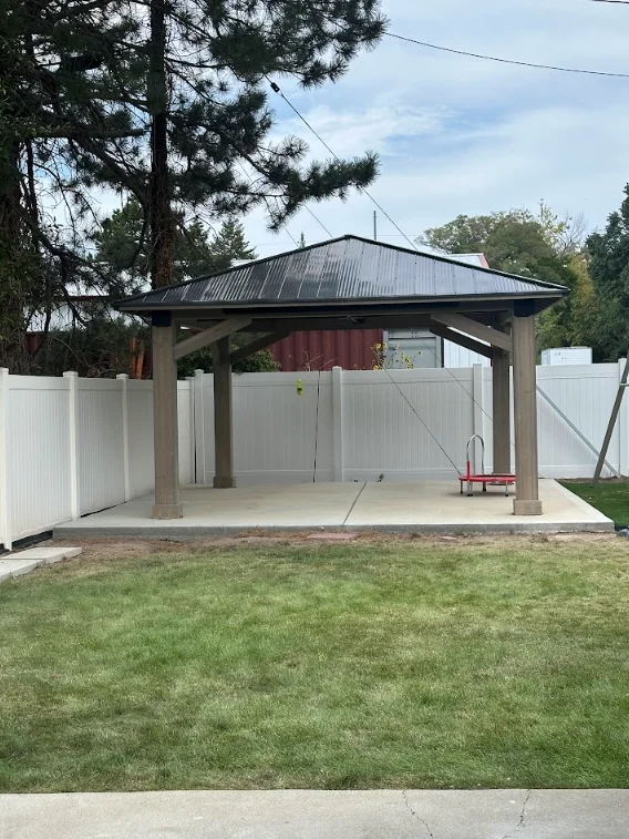 Wooden gazebo with black metal roof and concrete pad, white vinyl fence enclosure