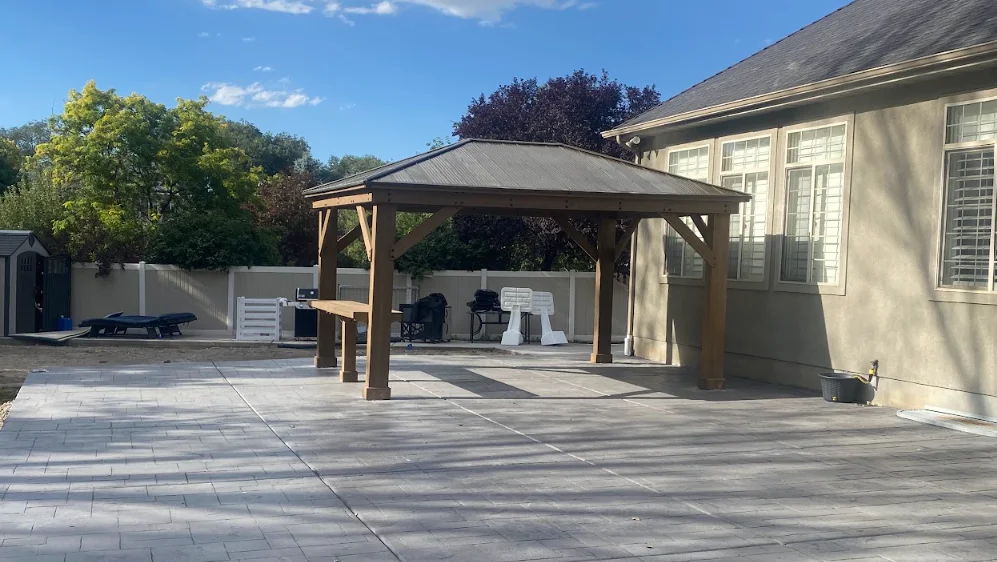 Wood gazebo with metal roof on gray paver patio beside residential home