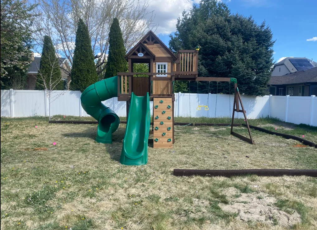 Wooden playset with green slides, climbing wall, playhouse, and swing set in fenced backyard