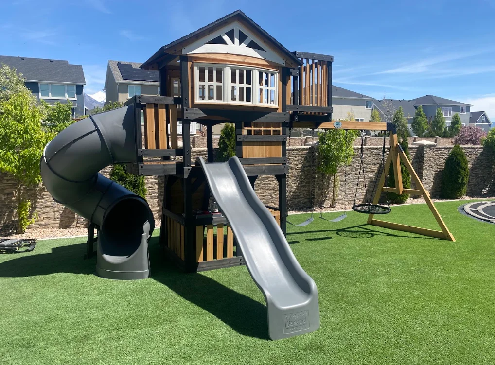Wooden playset with metal slides, tube slide, and swing set on synthetic grass with stone retaining wall